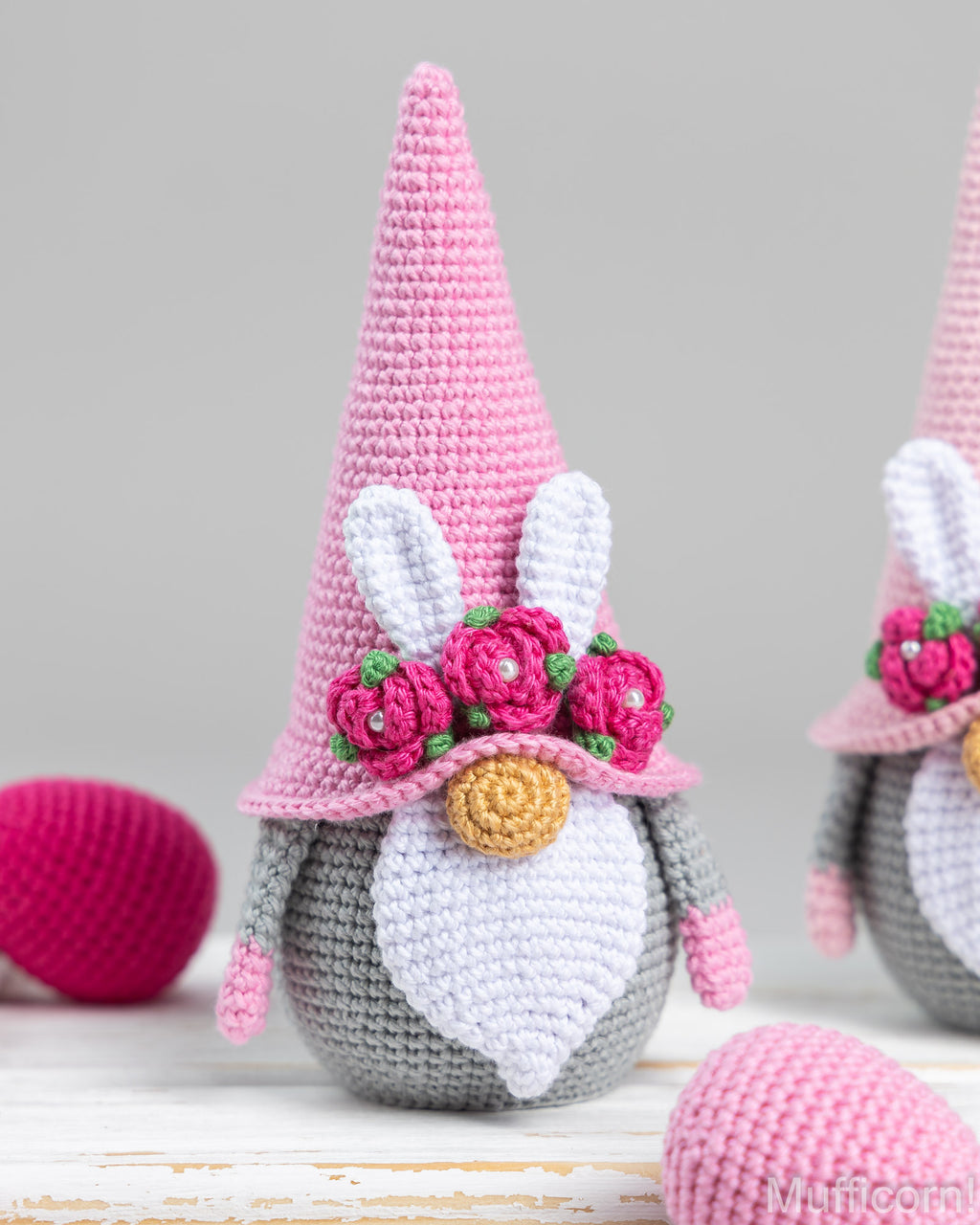 A crocheted bunny gnome decoration with a tall pink hat, white inner ears, a gray body, white beard, pink flowers with green leaves, and a gold nose, shown on a light surface with Easter eggs in the background.