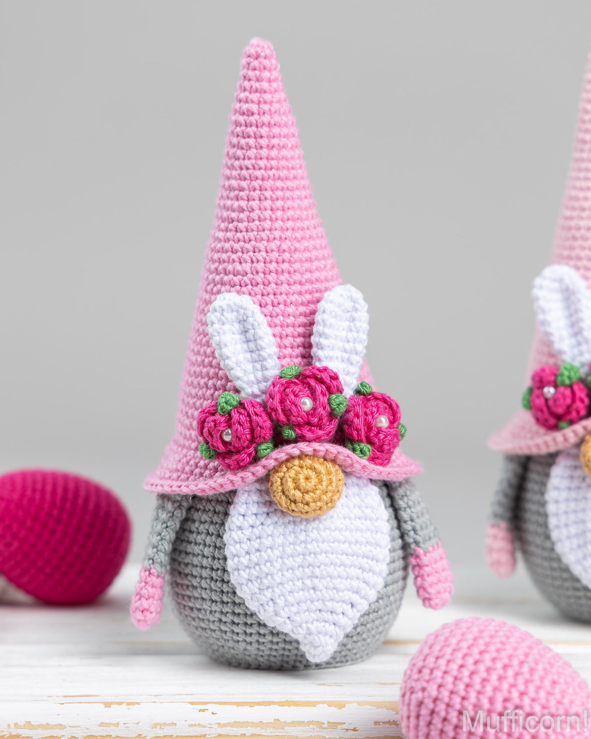 A crocheted bunny gnome decoration with a tall pink hat, white inner ears, a gray body, white beard, pink flowers with green leaves, and a gold nose, shown on a light surface with Easter eggs in the background.