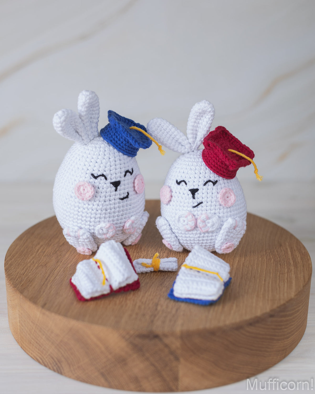 Two white crocheted bunny figures wearing graduation caps and gowns, displayed on a round wooden board against a light marble-like background.
