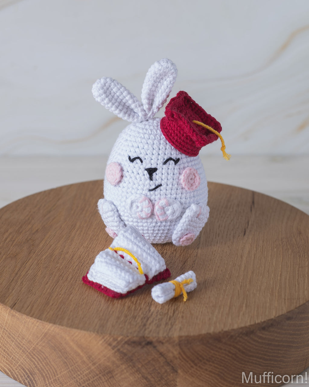 A crocheted white bunny plush toy wearing a red graduation cap and holding a diploma, displayed on a round wooden surface against a light marble background.