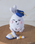 A crocheted white bunny amigurumi toy wearing a blue graduation cap with a yellow tassel, sitting on a round wooden surface. A matching blue and white diploma and scroll are placed in front.