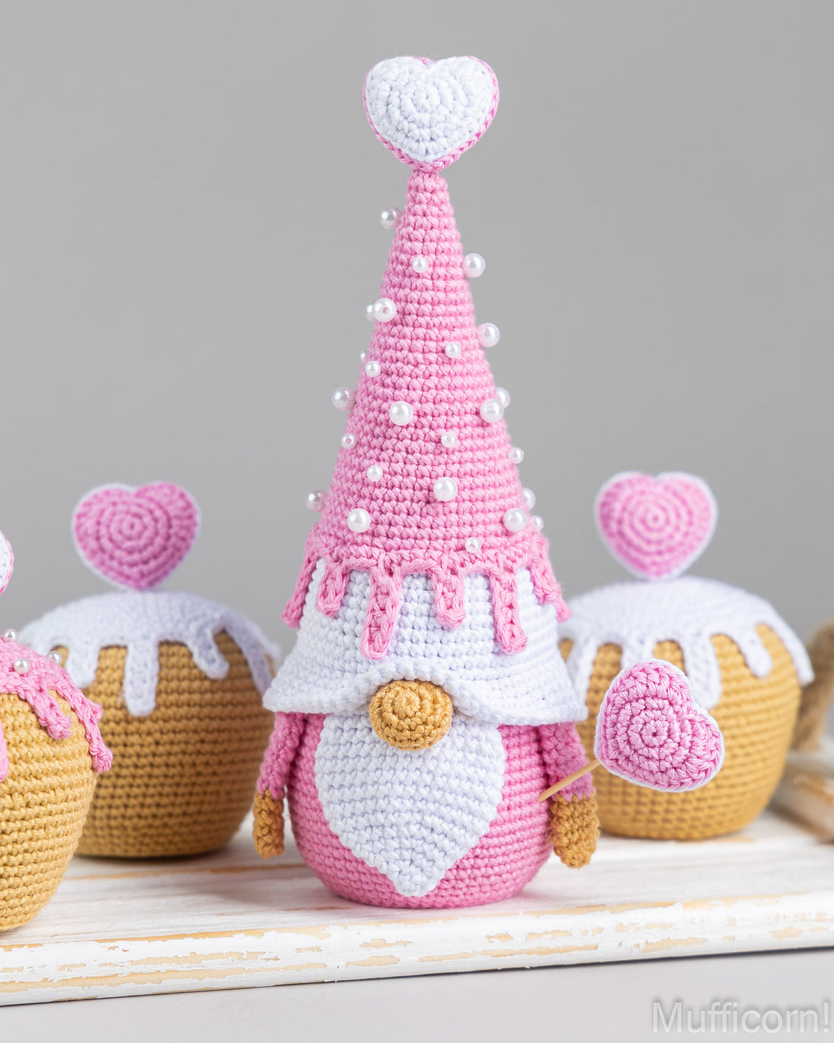 A crocheted Valentine's Day gnome decoration in pink and white stands on a white surface, surrounded by matching cupcake-shaped decorations with pink hearts on a soft gray background.