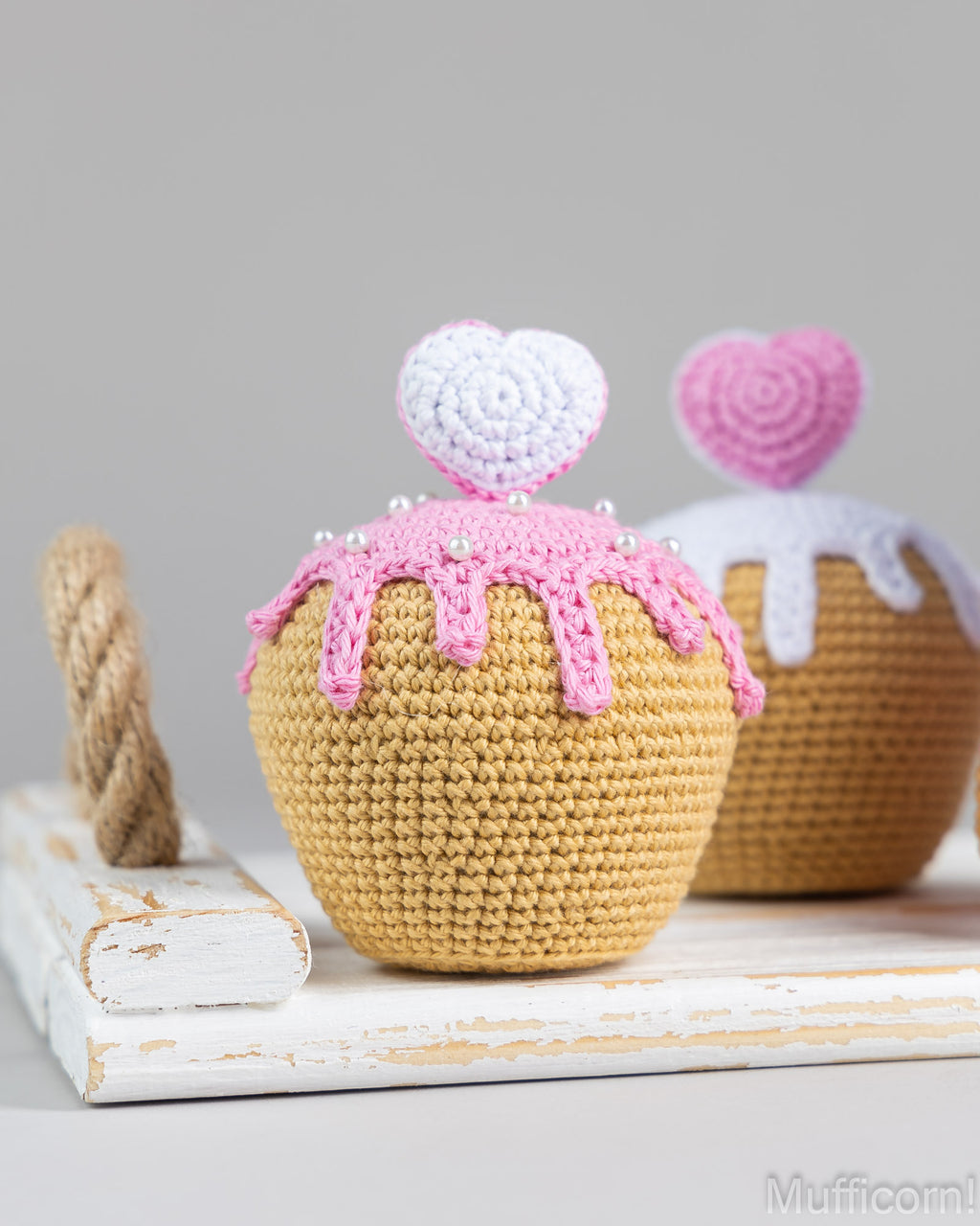 Two crocheted cupcake ornaments displayed on a white rustic board against a light gray background. The front cupcake is tan with pink frosting and a white-and-pink heart topper, while the rear cupcake is tan with white frosting and a pink heart topper.