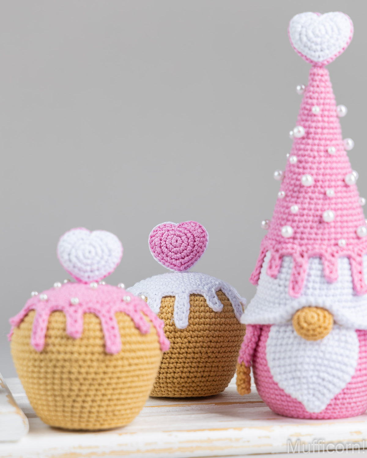 Three crocheted cupcake-shaped gnomes arranged on a white surface against a light gray background. The gnomes are pink, white, and yellow, with one tall pink gnome on the right and two shorter yellow and brown cupcake gnomes on the left.