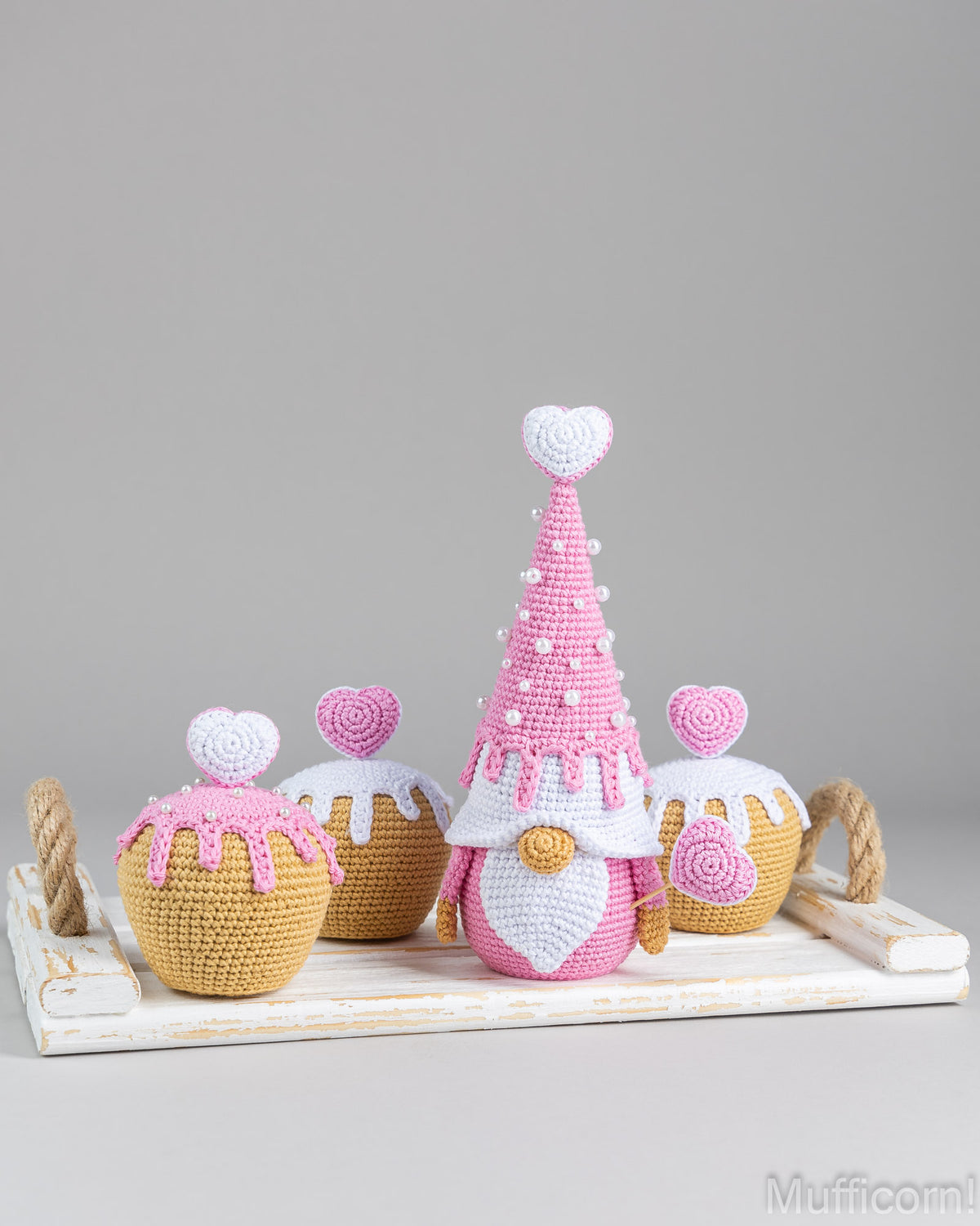 A crocheted gnome figurine styled like a cupcake, flanked by three smaller cupcake-like decorations on a white tray with rope handles, photographed against a neutral gray background.