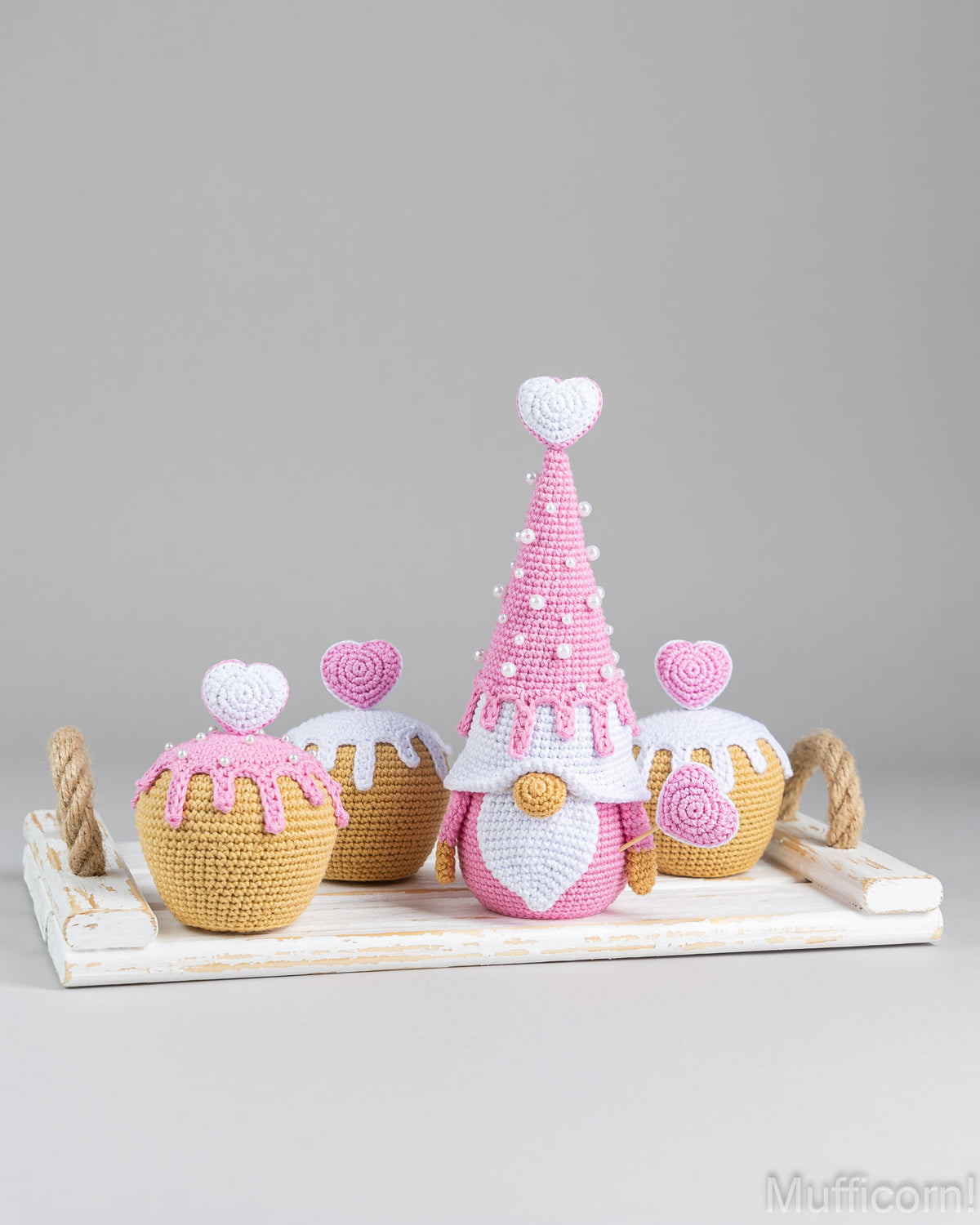 A crocheted gnomelike decoration with a pink and white body stands on a white tray beside three cupcake-shaped decorations, photographed against a plain light gray studio background.