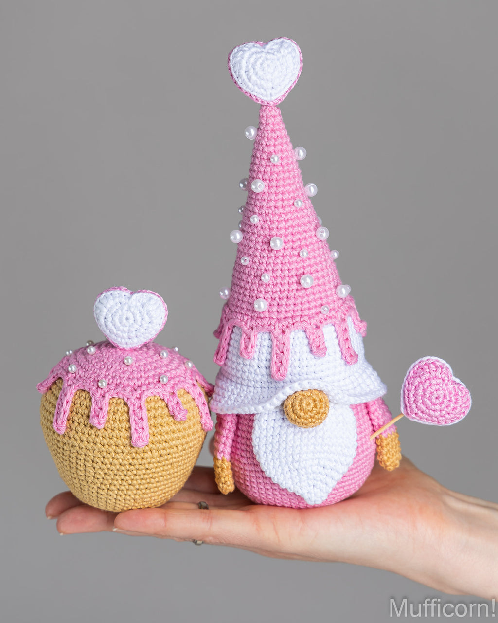 A hand holds two crocheted gnomes decorated like cupcakes. One gnome is tall and pink with a white beard, white hat, and pink hat trim, while the other is shorter and yellow with pink frosting details.