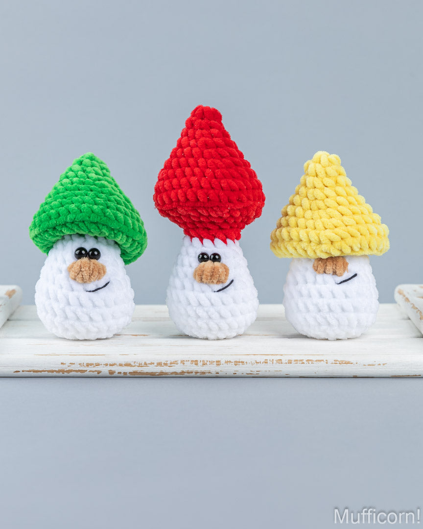 Three crocheted mushroom-shaped ornaments or small figurines standing on a white wooden surface against a light gray background. The left one has a green cap, the center one has a red cap, and the right one has a yellow cap.