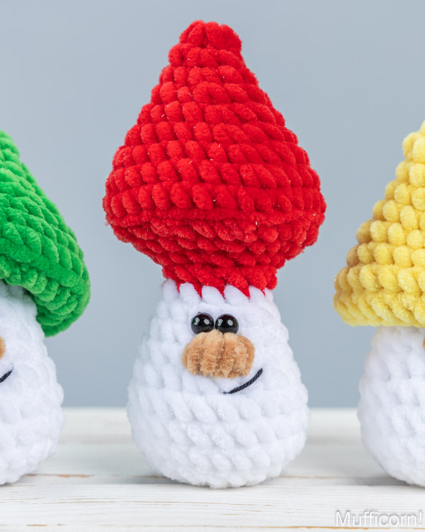 Three crocheted snowman ornaments displayed on a white wooden surface against a light gray background. The central snowman wears a textured red hat, with green and yellow hats partially visible on the left and right snowmen.