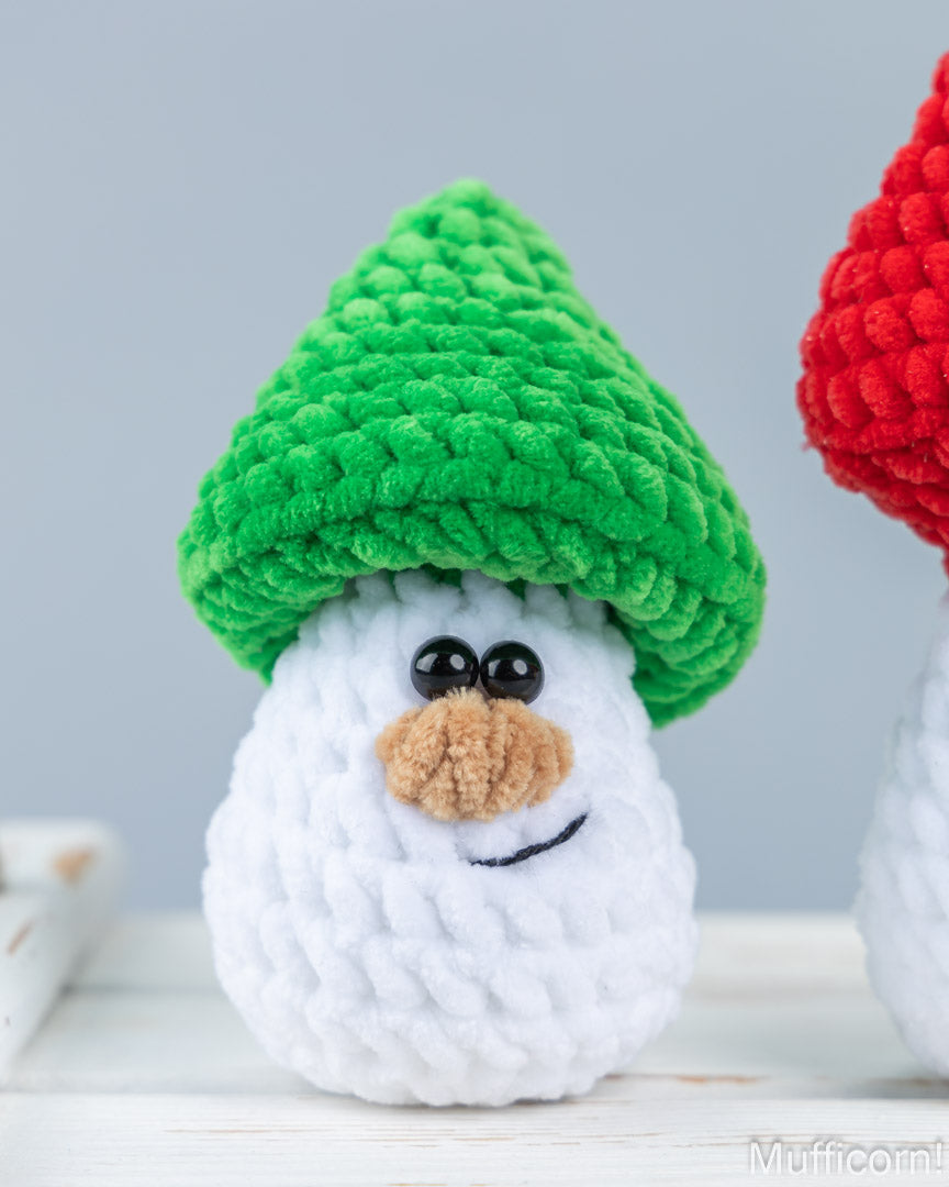 A crocheted snowman ornament wearing a green mushroom cap and a white body, photographed on a light surface against a pale gray background. Part of another snowman wearing a red cap is visible on the right edge.