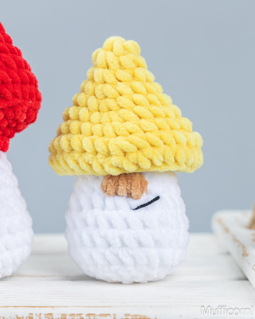 A close-up of a crocheted mushroom-shaped plush toy with a yellow textured cap, white body, and small brown face detail, sitting on a light surface against a pale blue background. Part of another red-and-white plush is visible at the left edge.