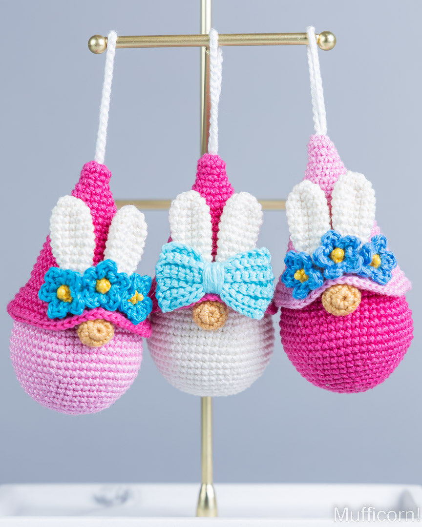 Three crocheted bunny egg ornaments hanging from a gold stand against a light gray background. The left ornament is pink and white with a blue bow and yellow flowers; the center ornament is white and pink with a light blue bow and yellow flowers; the right ornament is pink and white with a blue bow and yellow flowers.