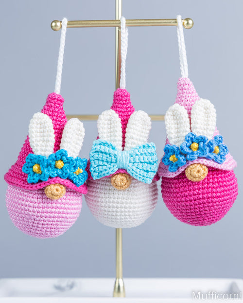 Three crocheted bunny egg ornaments hanging from a gold stand against a light gray background. The left ornament is pink and white with a blue bow and yellow flowers; the center ornament is white and pink with a light blue bow and yellow flowers; the right ornament is pink and white with a blue bow and yellow flowers.
