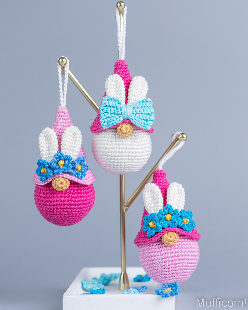 Three crocheted bunny egg ornaments displayed on a gold stand against a gray background. The center ornament is white with a pink top, a light blue bow, and blue flowers with yellow centers.