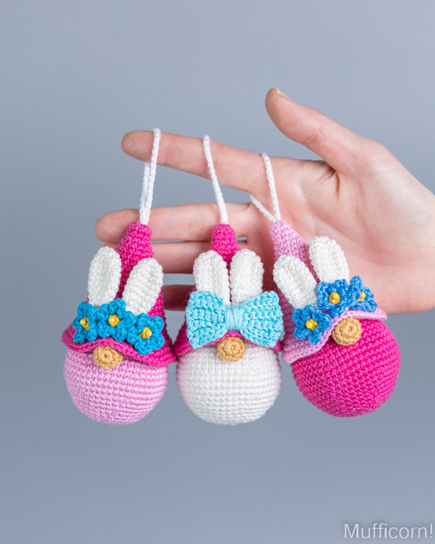 Three crocheted bunny egg ornaments are held in a person's hand against a plain gray background. The ornaments have white yarn ears, pink and white bodies, blue and pink bows, and small blue flower accents with yellow centers.