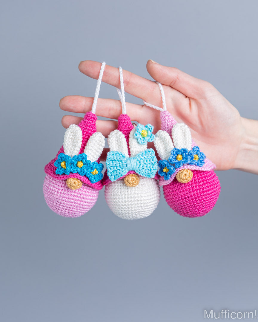 Three crocheted bunny egg ornaments in pink, white, and magenta, each with a blue flower and bow, held in a person's hand against a solid gray background.