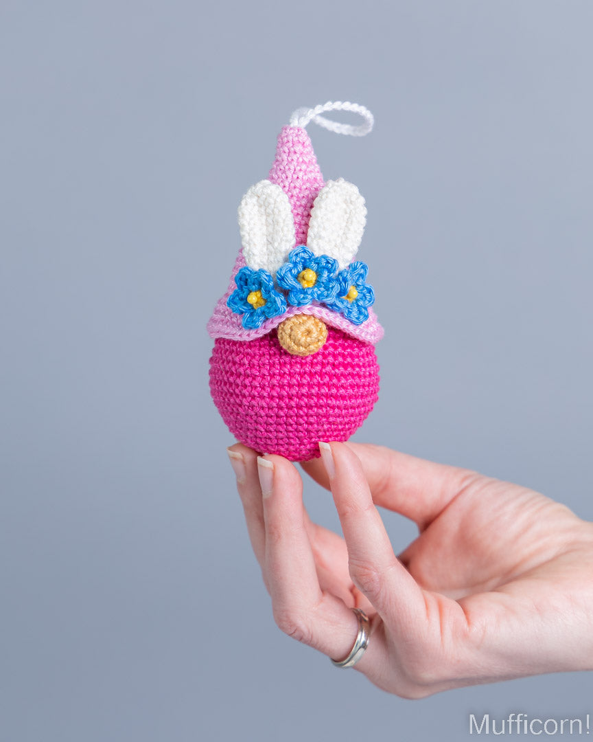 A hand holds a crocheted bunny ornament against a solid gray background. The ornament features a bright pink body, white bunny ears, blue flowers with yellow centers, and a small gold button on its face.