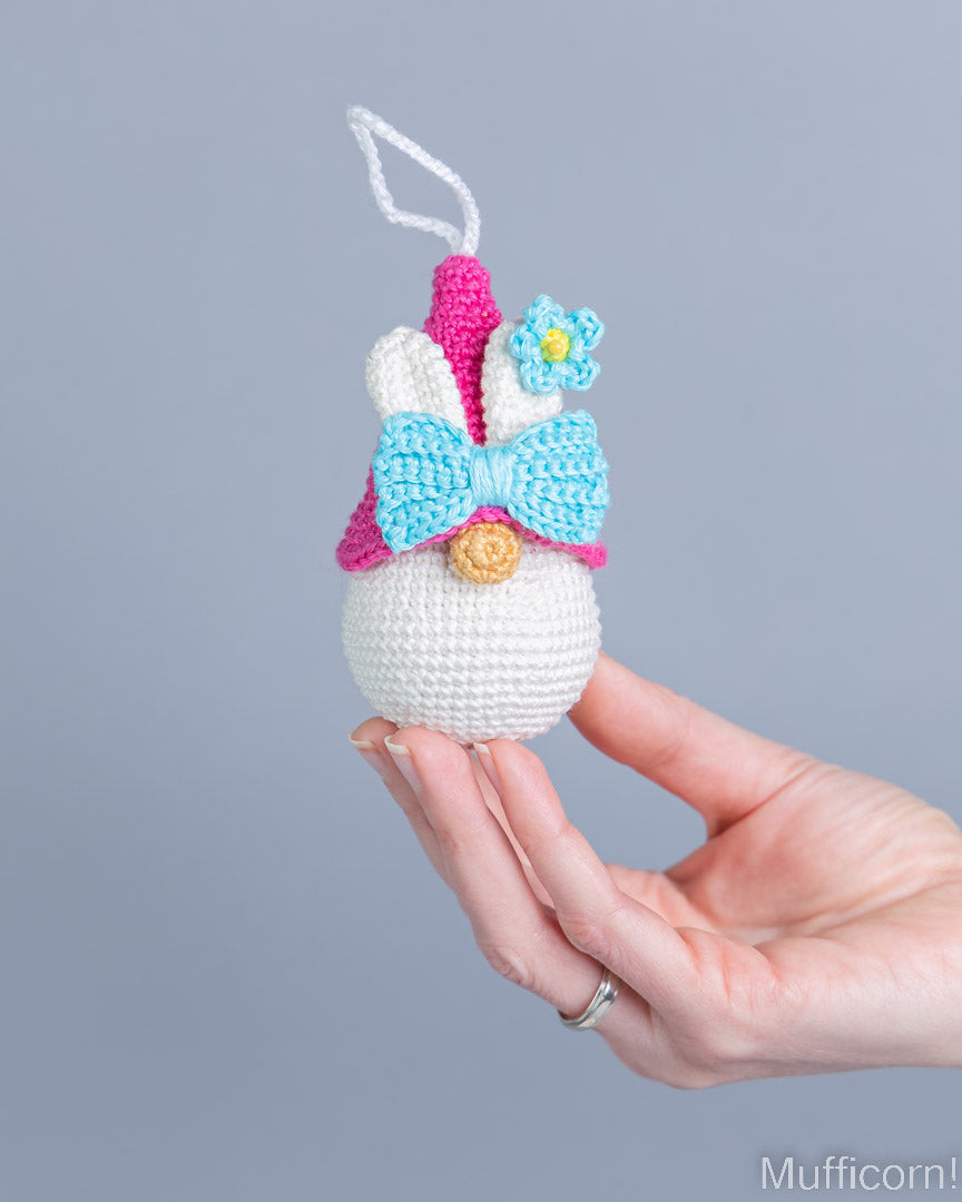 A hand holds a small crocheted bunny ornament against a plain gray background. The ornament is white with a pink and light blue bow, white ears, a pink nose, and a small blue flower accent.