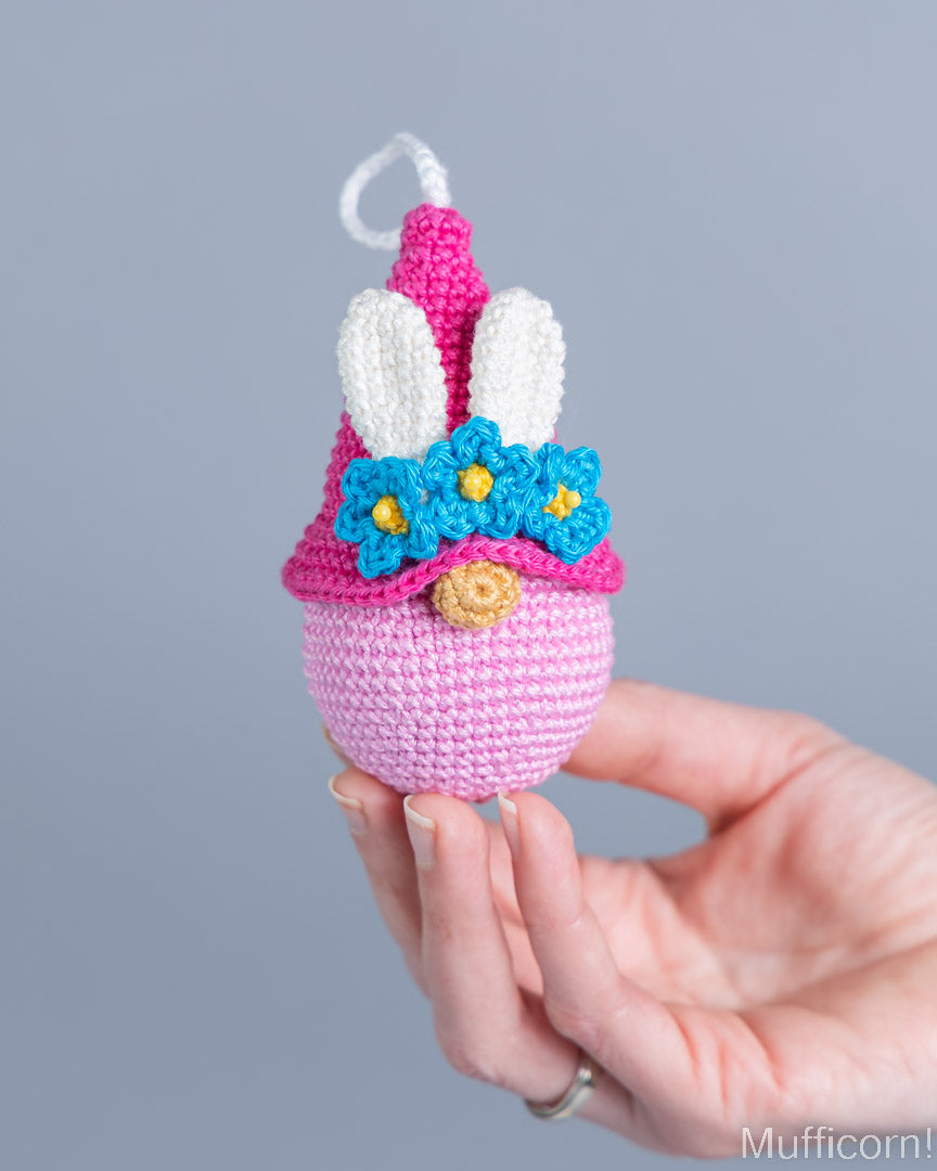A hand holds a small crocheted bunny-themed ornament or decoration against a plain light gray background. The item is primarily pink and lavender, with white bunny ears, blue flower accents, yellow centers, and a small gold button detail.