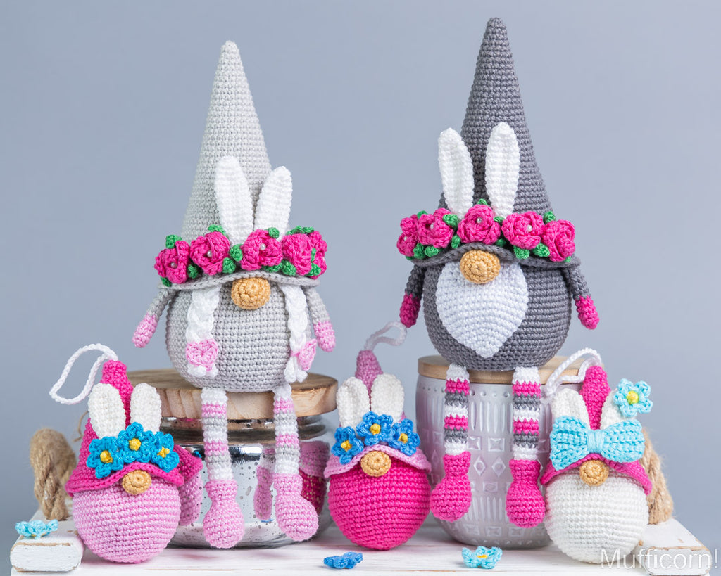 A set of handmade crocheted Easter-themed items, including two gnome figures, pink and white bunny eggs, and small flower decorations, arranged on a white surface against a solid gray background.