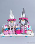 A collection of crocheted Easter-themed gnome and bunny decorations displayed on a white wooden shelf against a solid gray studio background. The set includes two tall gray gnomes with pink flower crowns and white beards, several smaller pink, white, and blue bunnies, and matching pink and white striped socks.