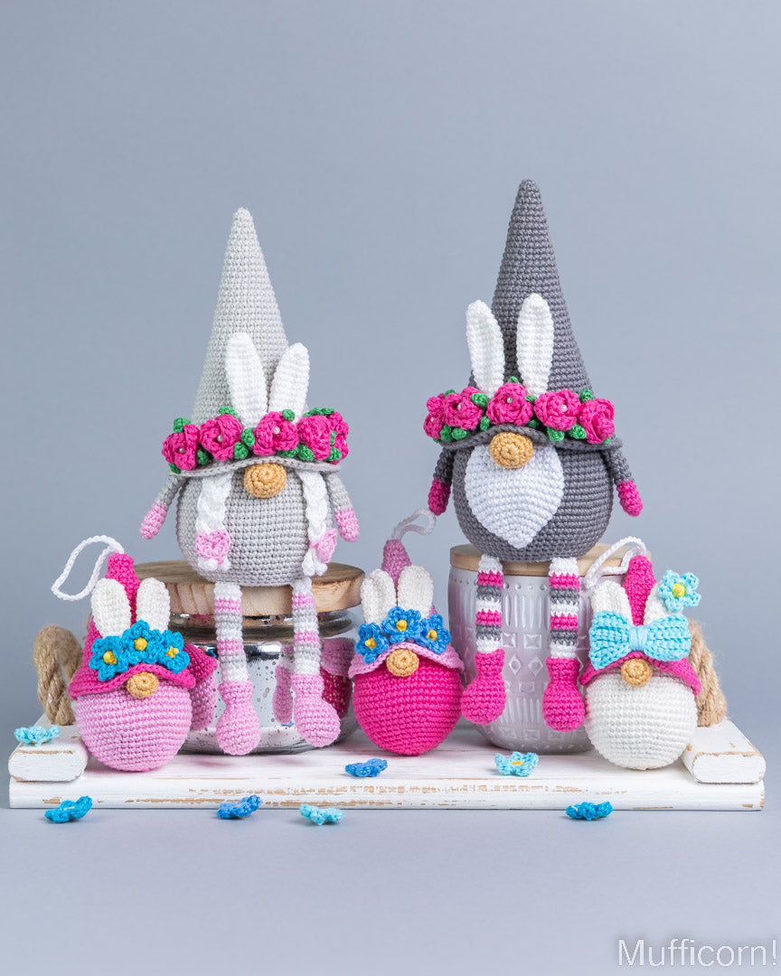 A collection of crocheted Easter-themed gnome and bunny decorations displayed on a white wooden shelf against a solid gray studio background. The set includes two tall gray gnomes with pink flower crowns and white beards, several smaller pink, white, and blue bunnies, and matching pink and white striped socks.