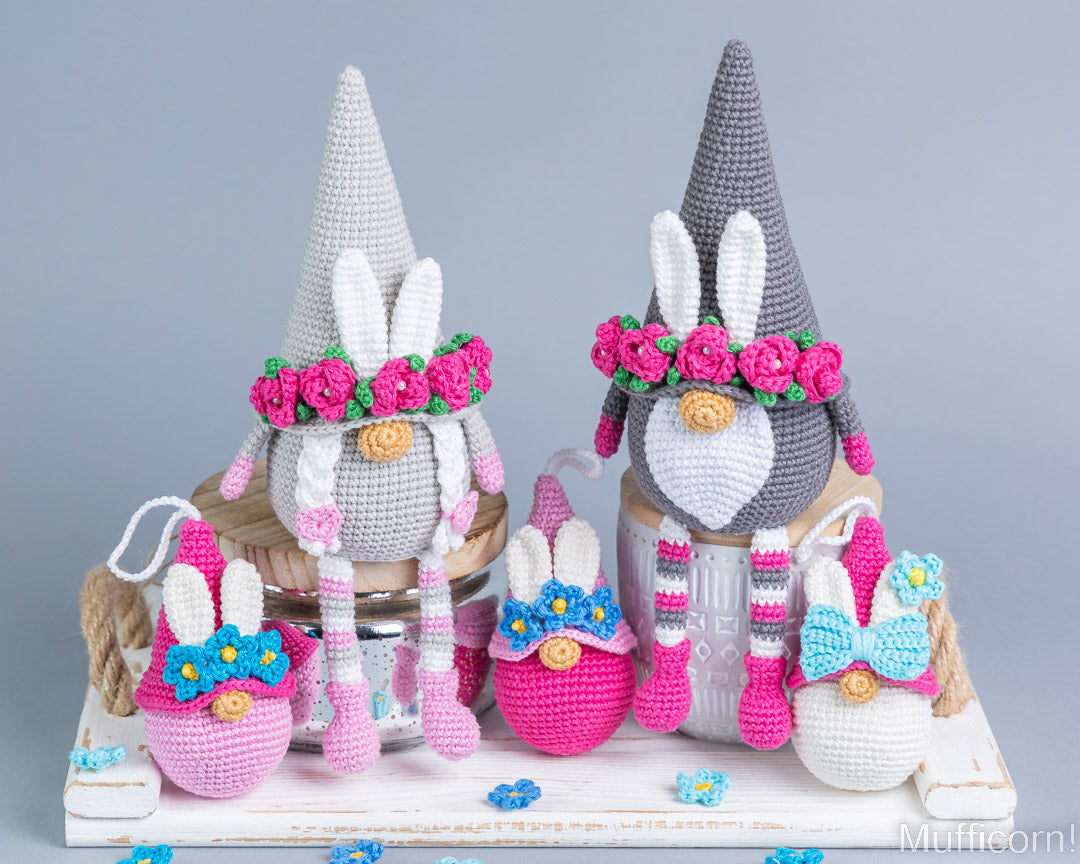 A collection of crocheted Easter-themed decorative items, including two gnome figures with tall pointed hats, pink flower crowns, bunny ears, and colorful egg-shaped ornaments, arranged on a white wooden surface against a light gray background.