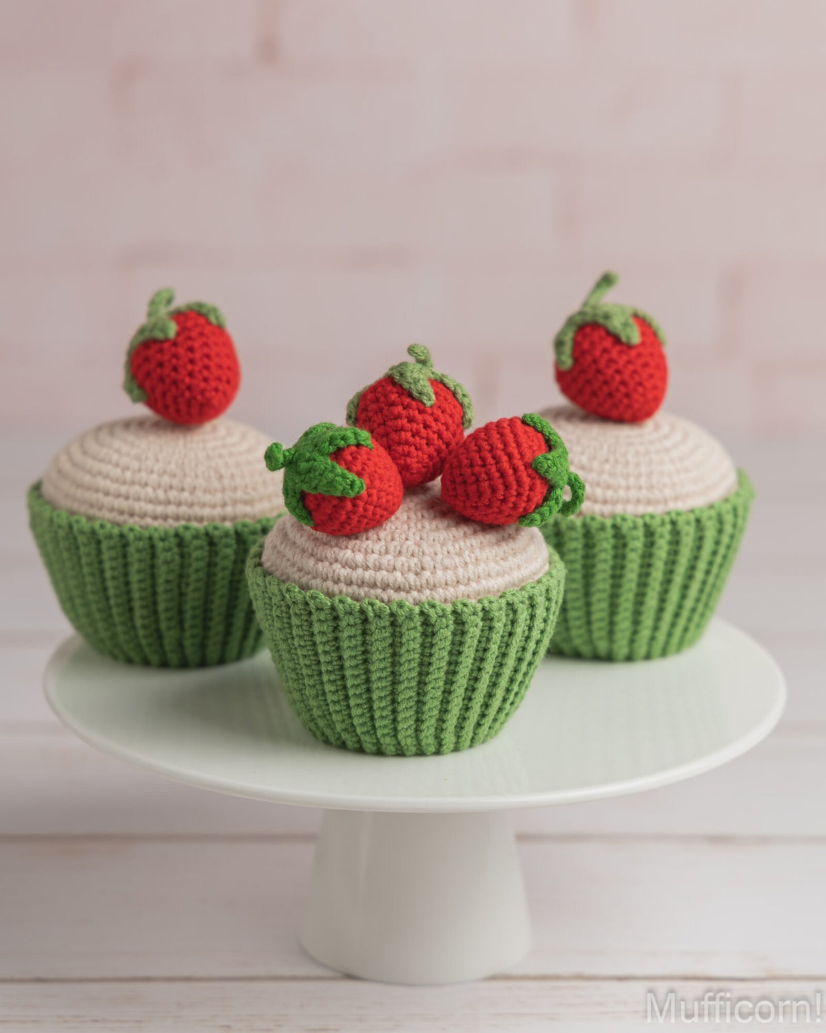 Three crocheted strawberry cupcakes displayed on a white cake stand against a soft pink brick wall background.