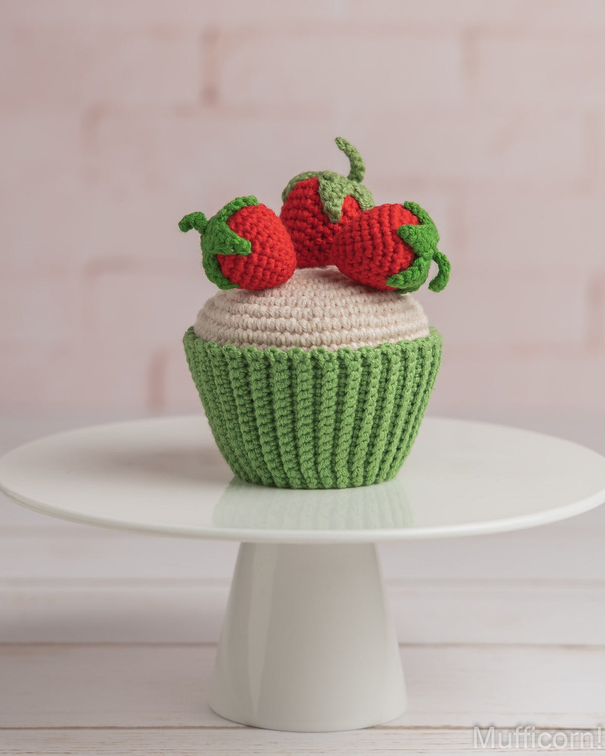 A crocheted cupcake decoration or toy with a green textured wrapper, cream-colored frosting, and three red strawberries on top, displayed on a white pedestal stand against a soft pink brick wall background.