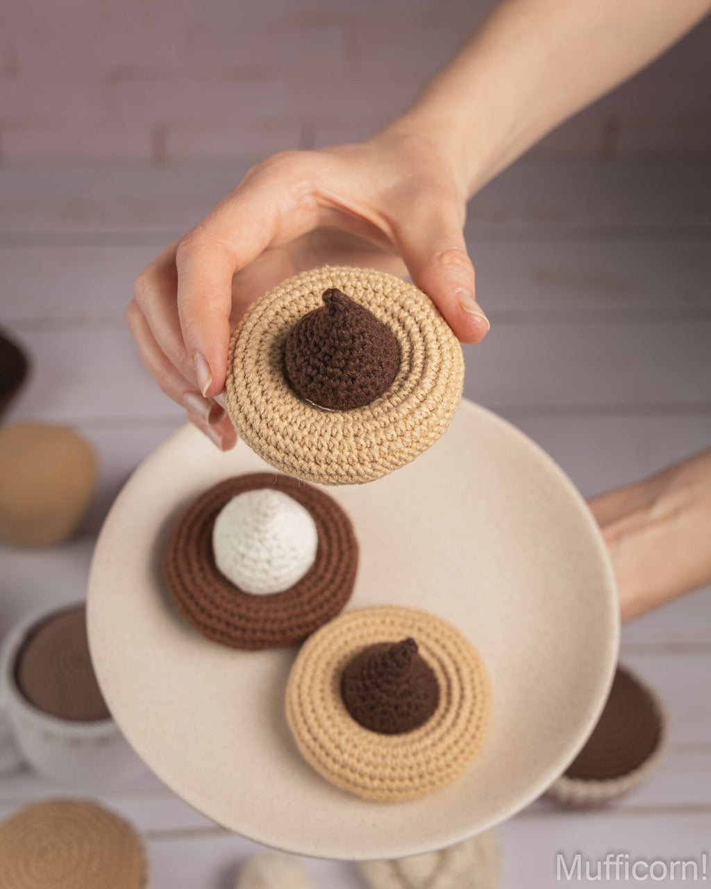 A hand holds a crocheted acorn-shaped item above a plate with similar acorns, photographed in a softly styled kitchen-like setting. The product appears to be a handmade crochet accessory or decoration in beige and brown tones.