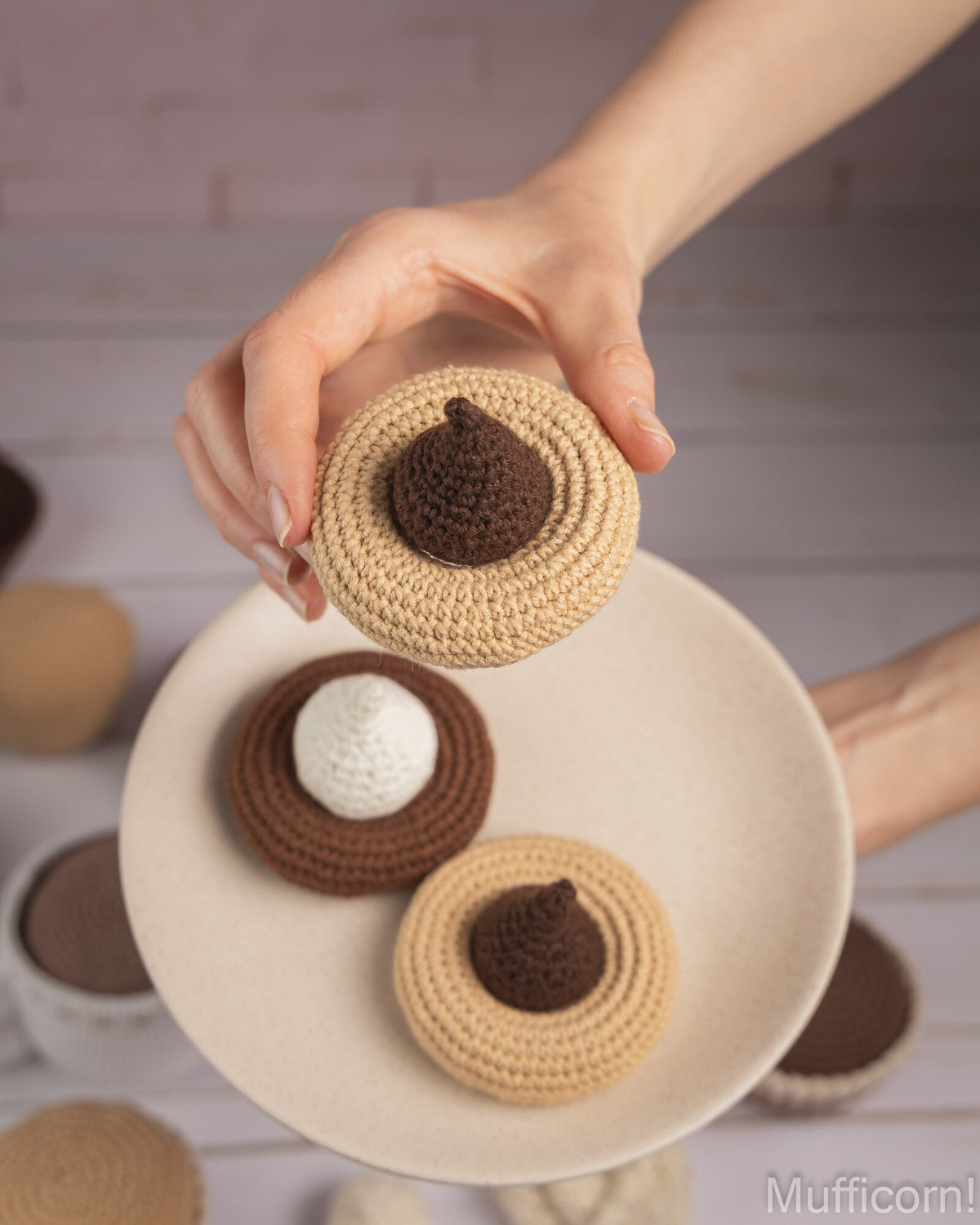 A hand holds a crocheted acorn-shaped item above a plate with similar acorns, photographed in a softly styled kitchen-like setting. The product appears to be a handmade crochet accessory or decoration in beige and brown tones.