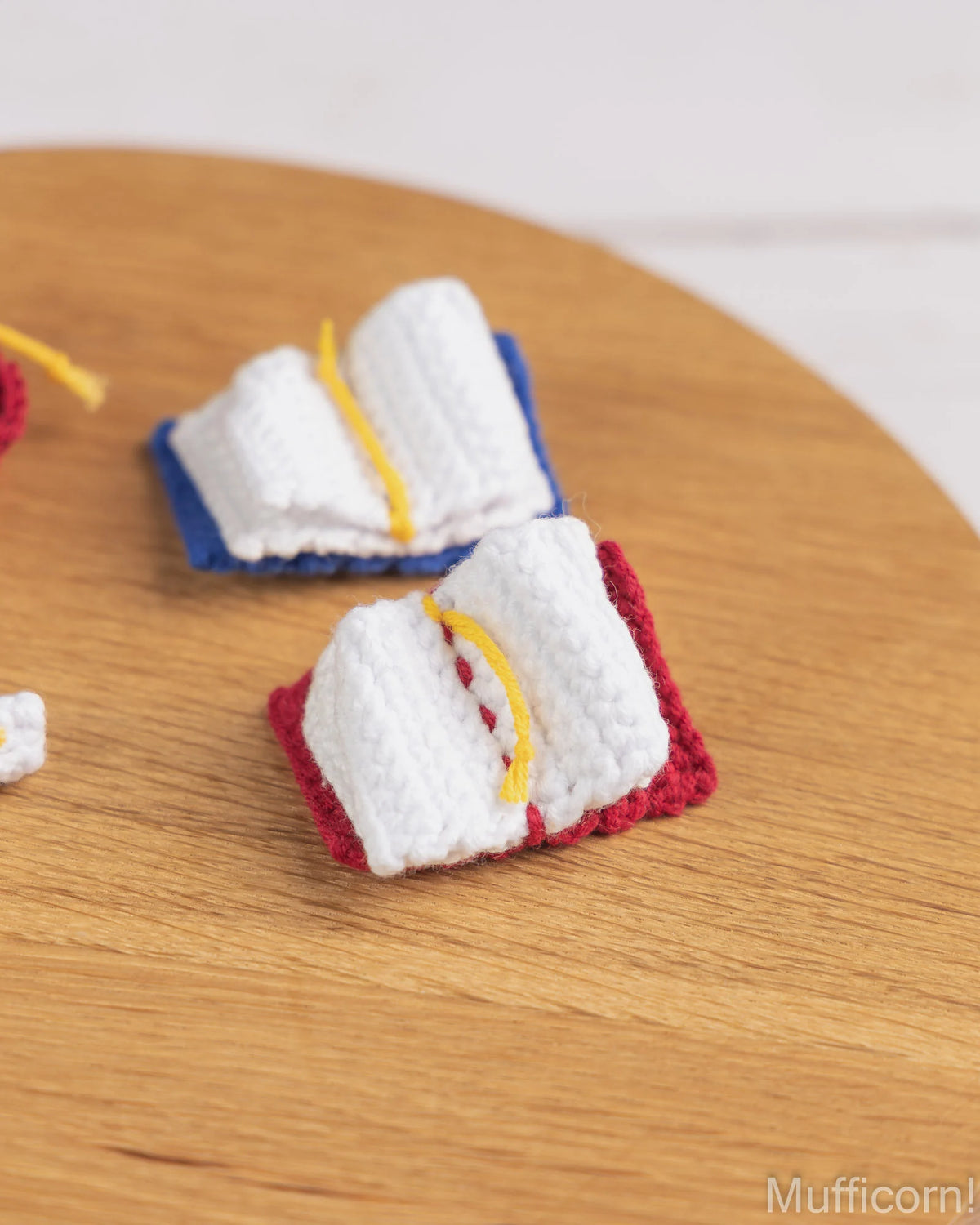 Two miniature white crochet book charms with red and blue bases and yellow thread details, displayed on a light wood surface.