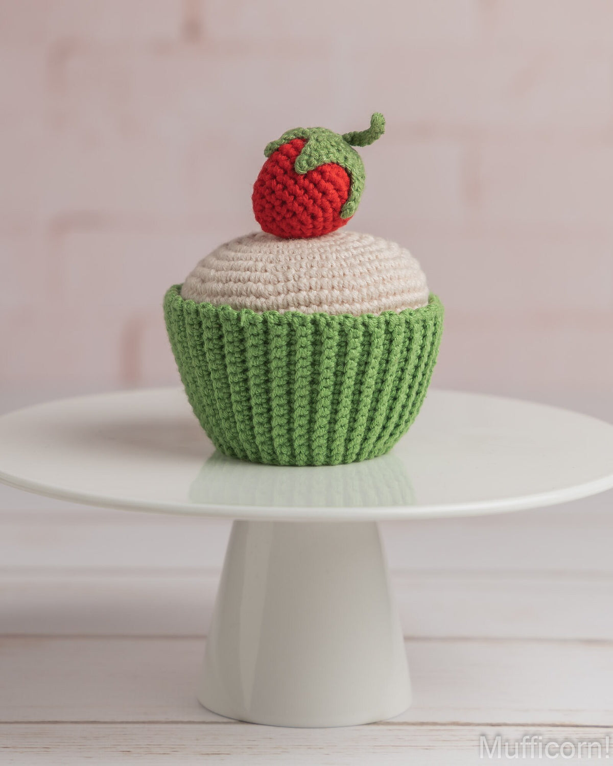 A crocheted cupcake decoration or toy with a green textured wrapper, cream-colored frosting, and a red strawberry on top, displayed on a white round pedestal against a soft pink background.