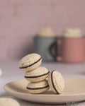 Three crocheted macarons in cream and brown, stacked on a beige plate with a soft pink background and blurred mugs.
