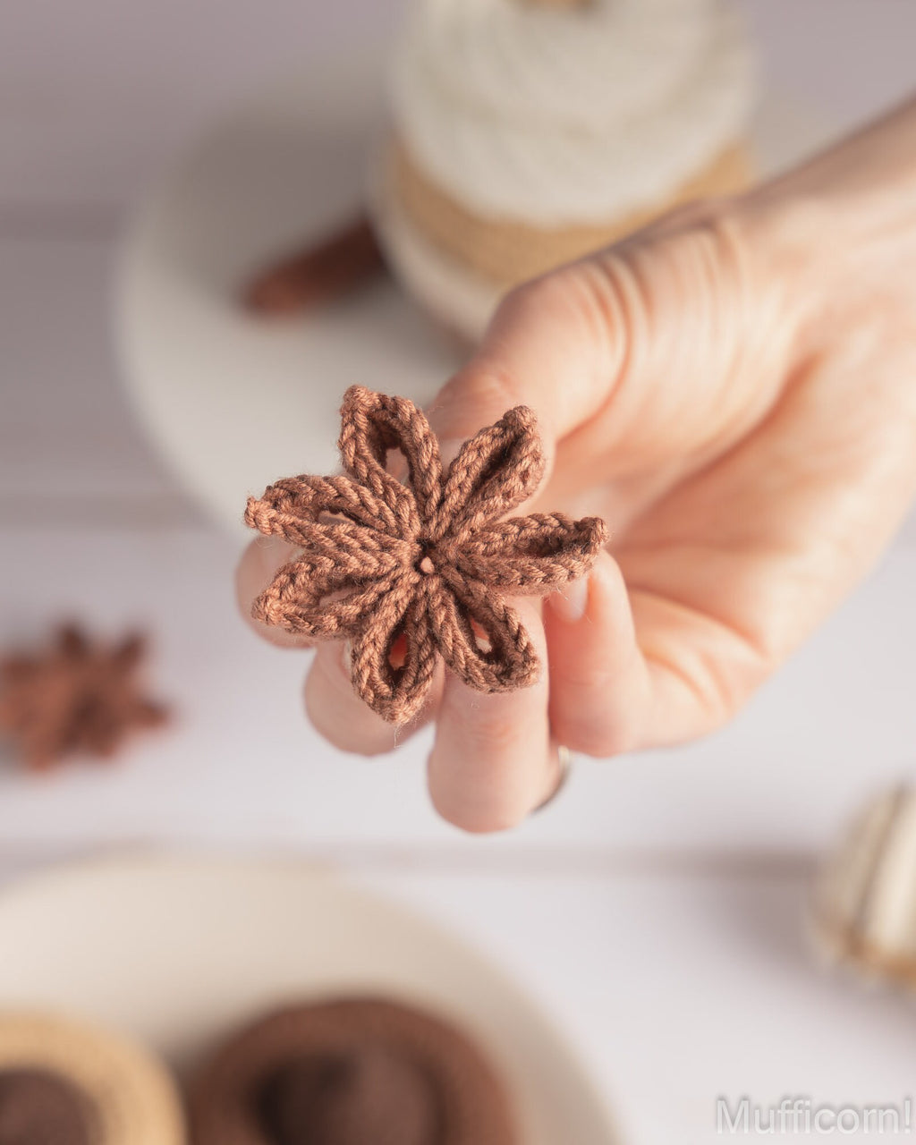 A hand holding a small crocheted brown star anise ornament, with blurred star anise and a spool of thread in the background on a white surface.