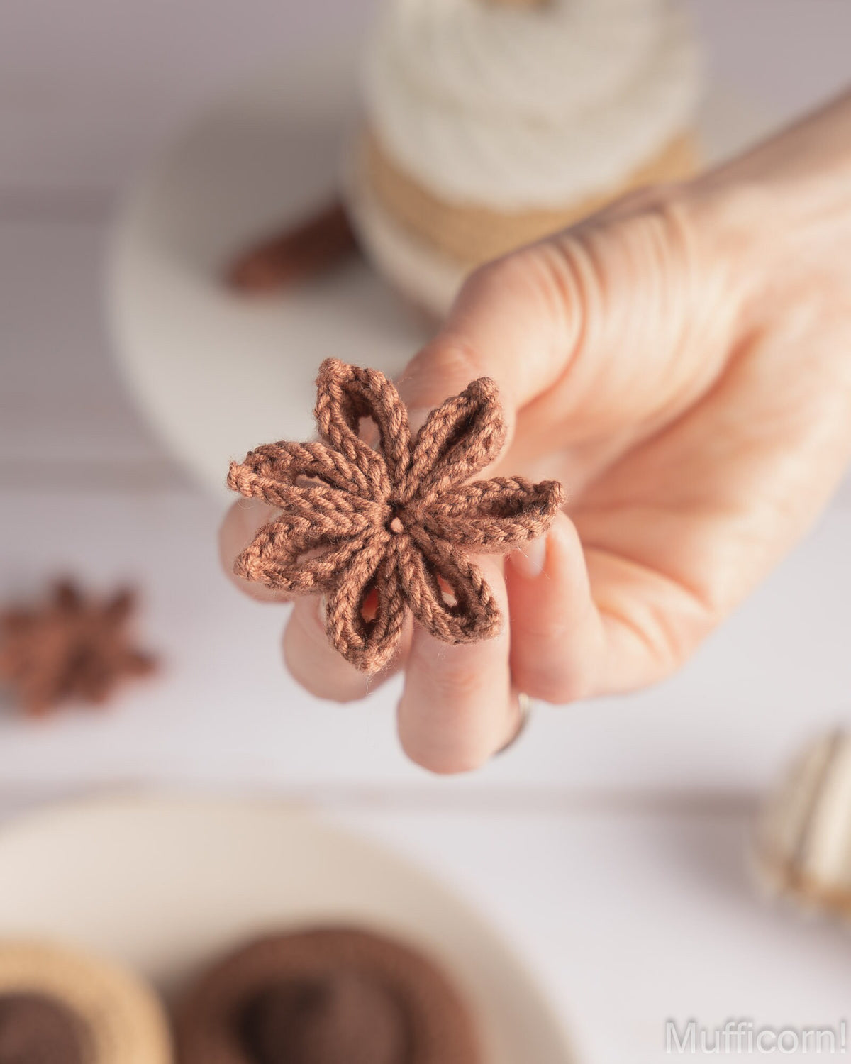 A hand holding a small crocheted brown star anise ornament, with blurred star anise and a spool of thread in the background on a white surface.