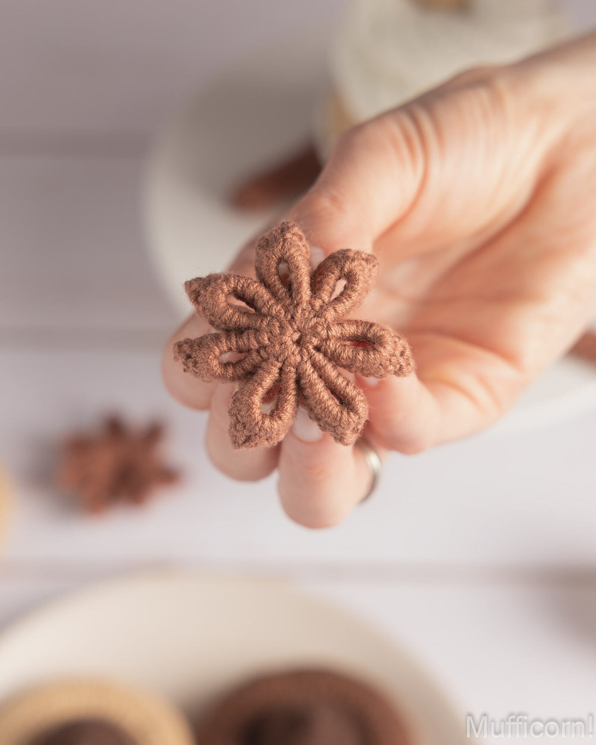 A hand holding a small, textured brown star-shaped pastry or cookie against a softly blurred light background with more pastries visible on plates.
