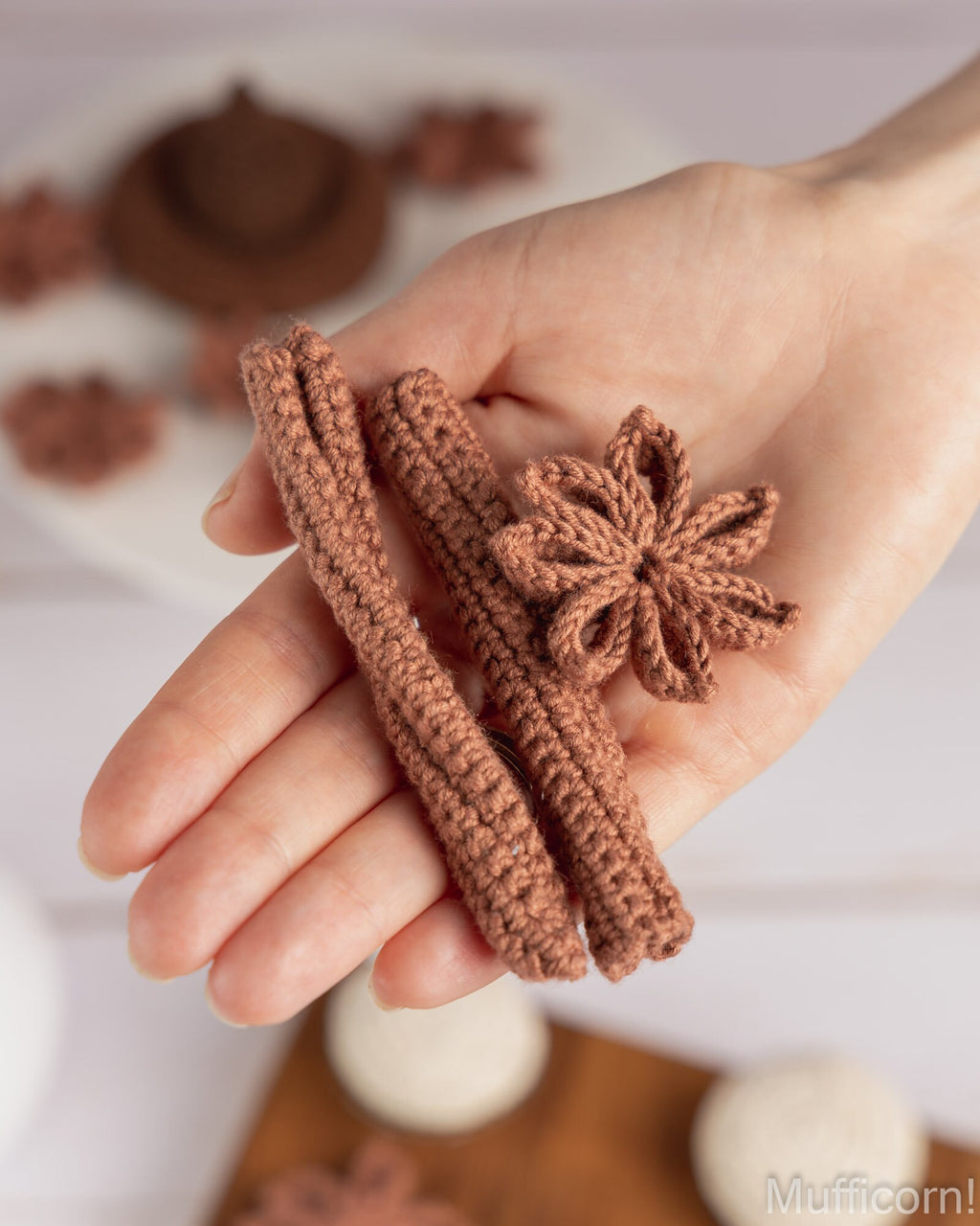 A hand holds a small crocheted set consisting of two cinnamon stick-shaped pieces and a flower-shaped piece in a warm brown color, with a softly blurred background.