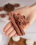 A hand holds a small crocheted set consisting of two cinnamon stick-shaped pieces and a flower-shaped piece in a warm brown color, with a softly blurred background.