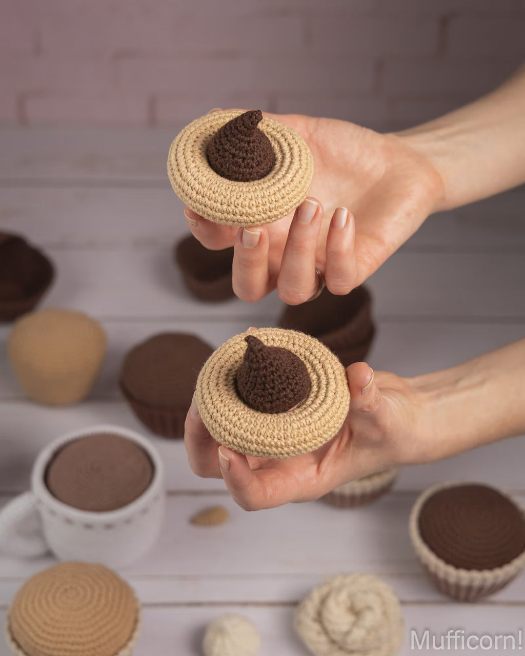 A person's hands holding two crocheted cupcake covers shaped like chocolate-covered pastries, with more similar cupcake covers scattered on a light wooden surface against a pale pink brick wall background.