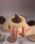 A hand holds a small crocheted chocolate-hershey's-style treat with a beige wrapper and dark brown center, against a softly blurred kitchen background with more similar treats.