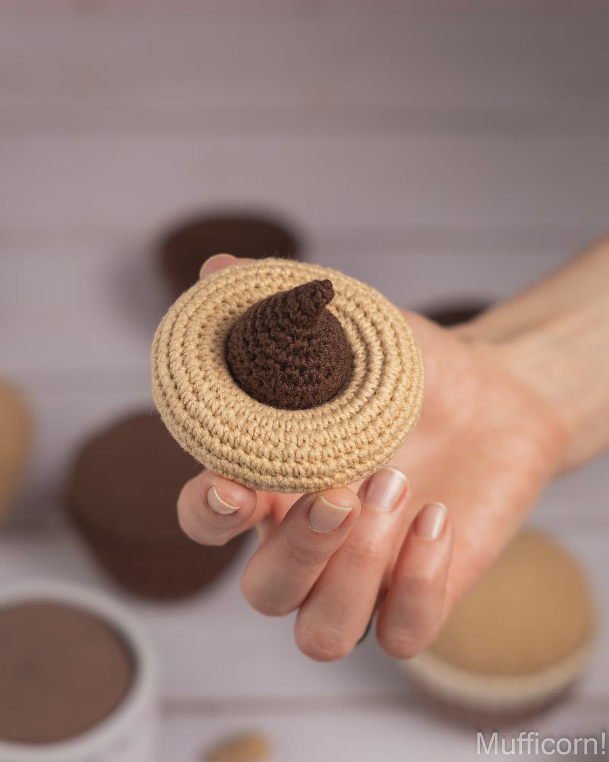 A hand holding a crocheted muffin in beige with a dark brown chocolate-look center, with more muffins blurred in the background.