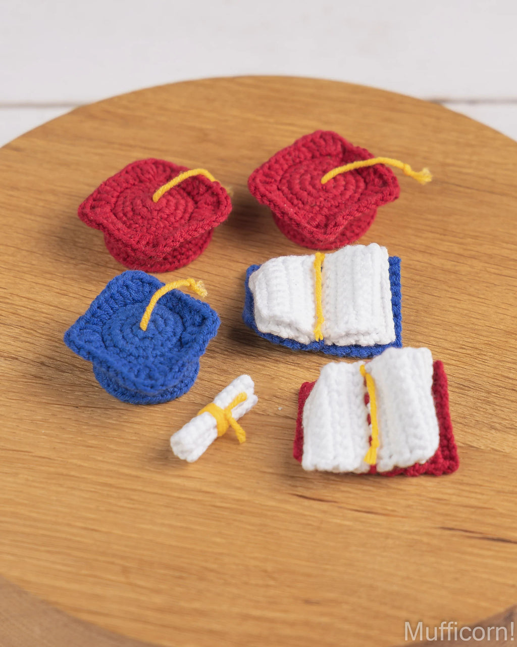 A set of miniature crochet graduation-themed accessories, including two red mortarboard caps, one blue cap, two white open books with red and blue covers, and a small white diploma scroll with a yellow tie, arranged on a round wooden surface.