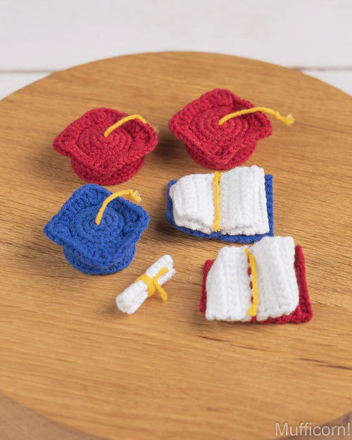 A set of miniature crochet graduation-themed accessories, including two red mortarboard caps, one blue cap, two white open books with red and blue covers, and a small white diploma scroll with a yellow tie, arranged on a round wooden surface.