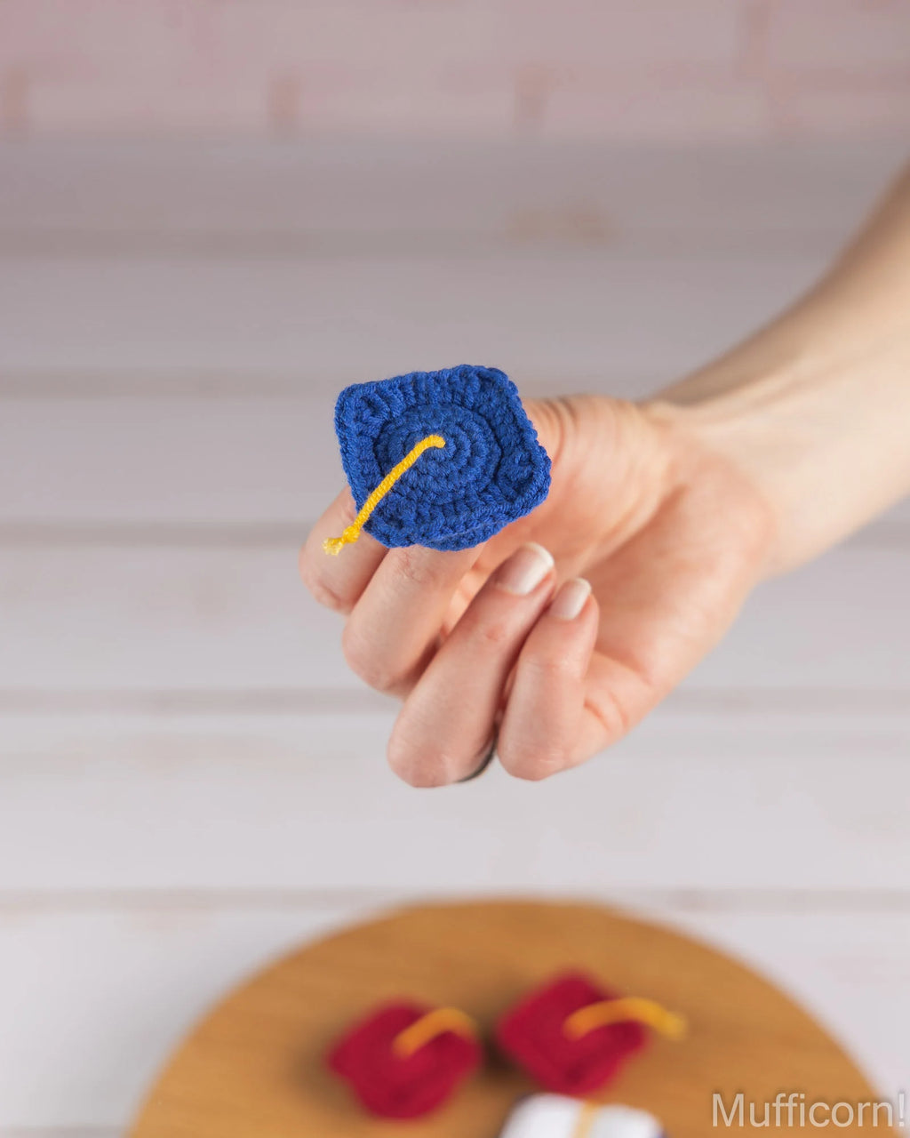 A hand holds a small crocheted blue graduation cap with a yellow tassel against a light wood background, with more small crocheted items blurred in the foreground.