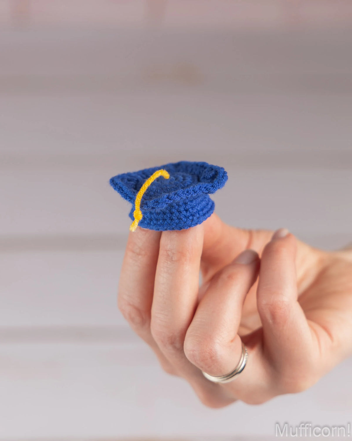 A miniature blue crochet graduation cap ring worn on a person's finger, featuring a yellow tassel. The hand is shown against a soft, blurred light gray background.