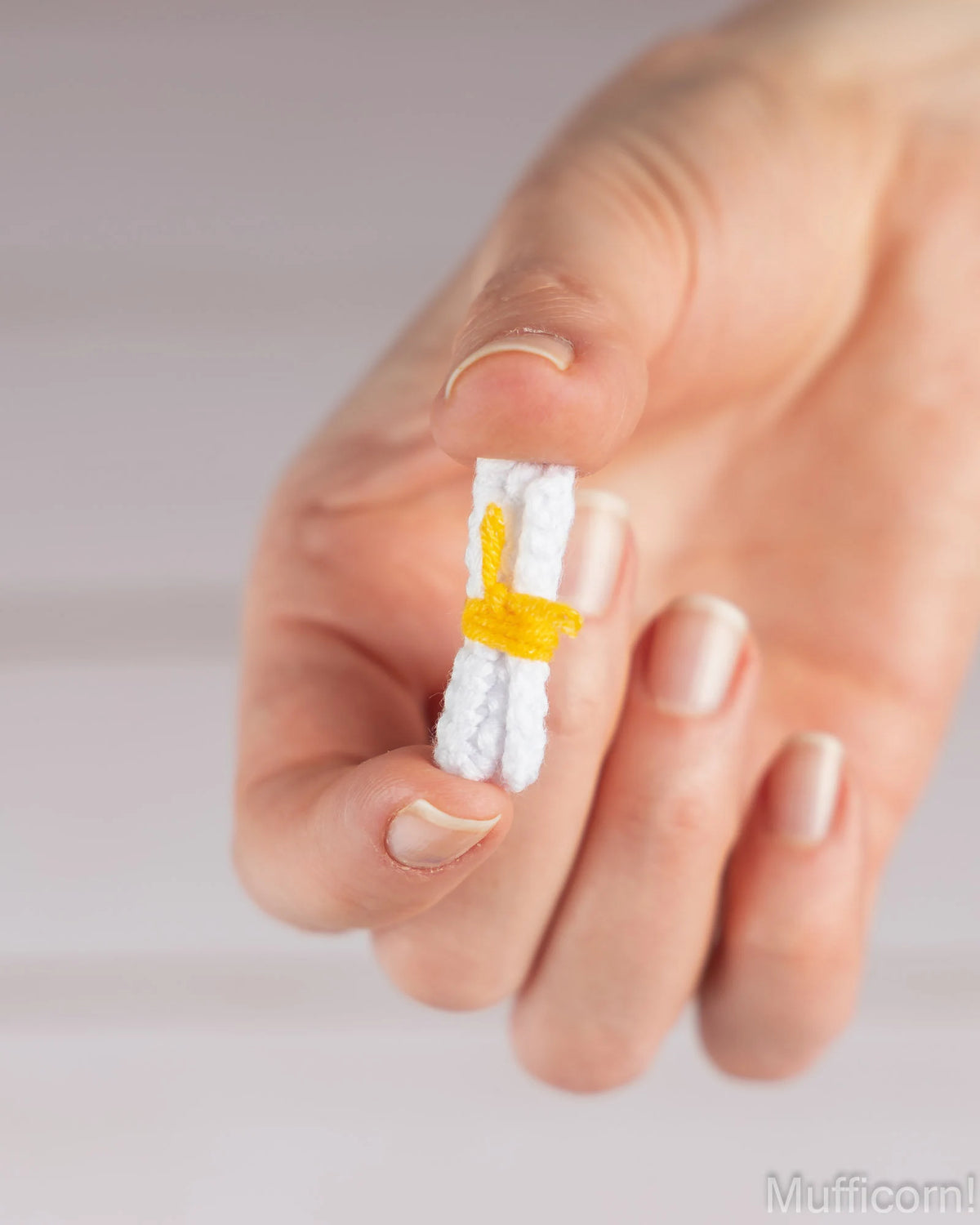 A close-up of a hand holding a small white novelty item wrapped with a yellow band, photographed against a soft light gray background.