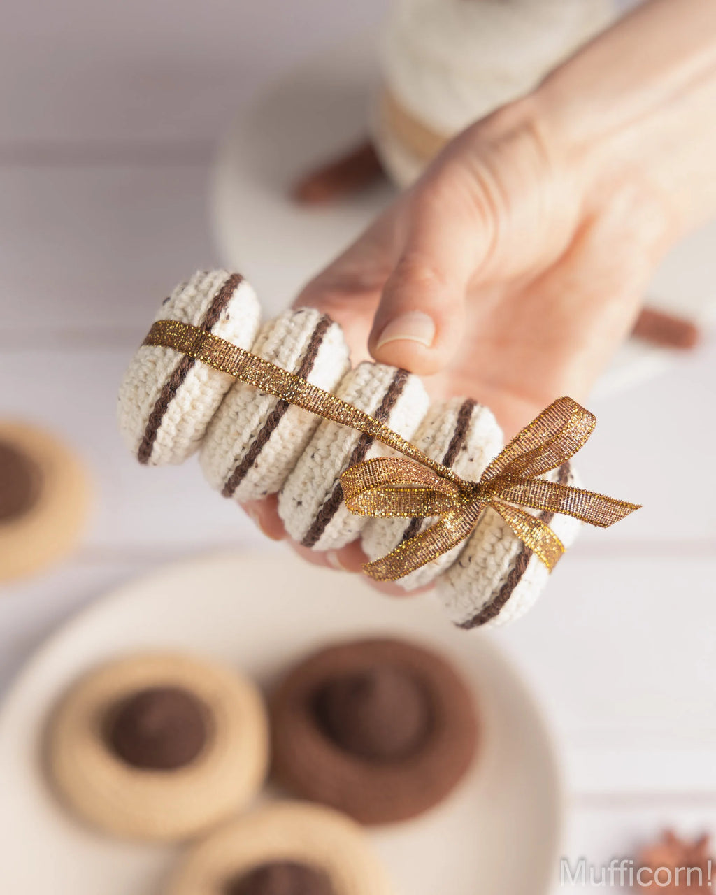 A hand holds a small decorative macarons.  Blurred cookies and plates appear in the background on a light surface.