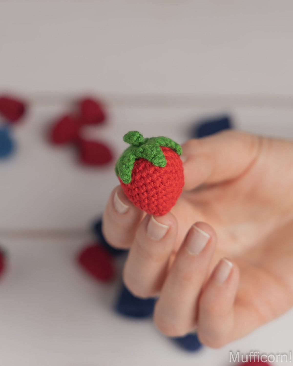 A hand holding a small crocheted strawberry amigurumi toy, with other colorful strawberry toys blurred in the background on a light surface.