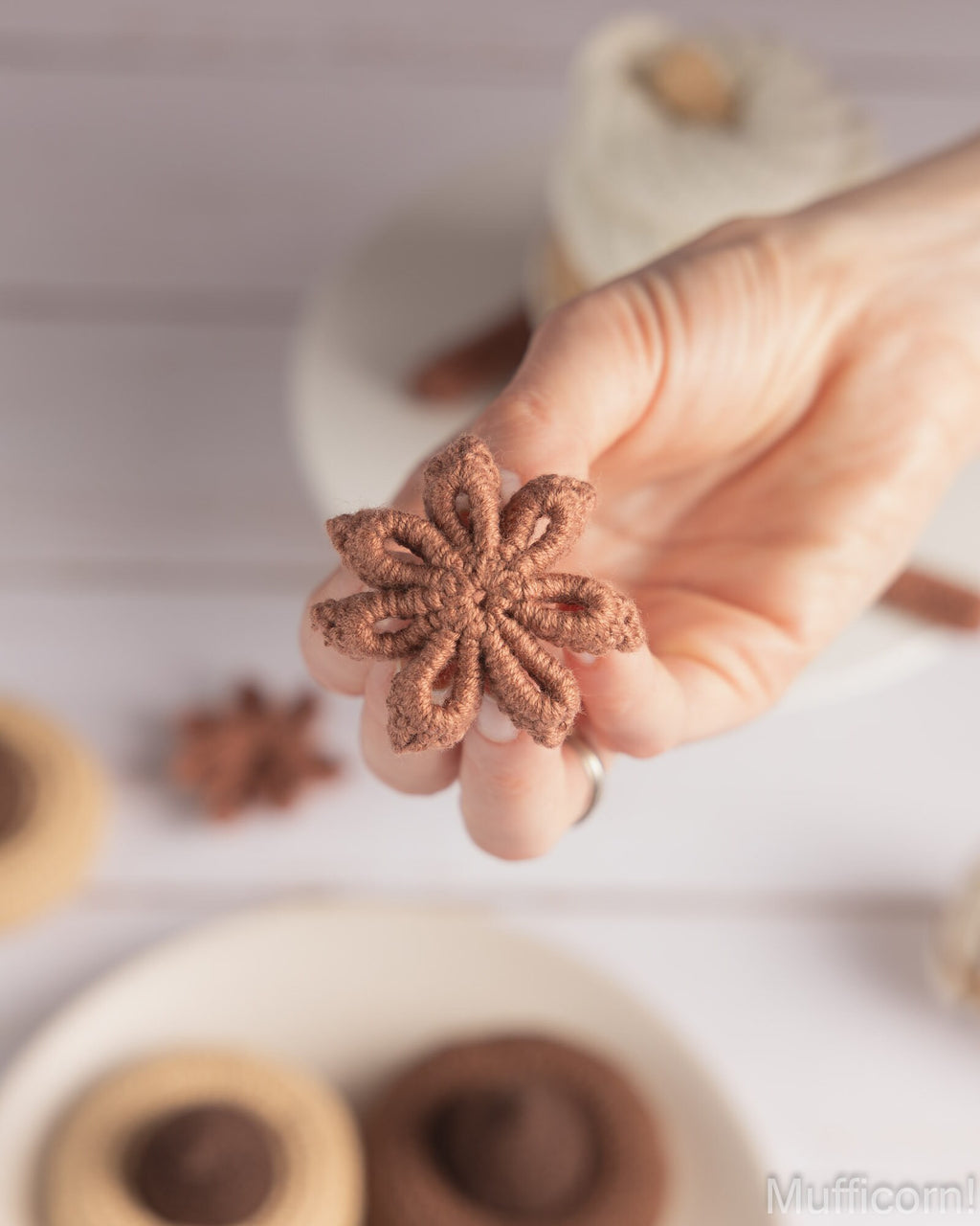 A hand holds a small, textured brown star-shaped cookie against a softly blurred background of white plates and more cookies.
